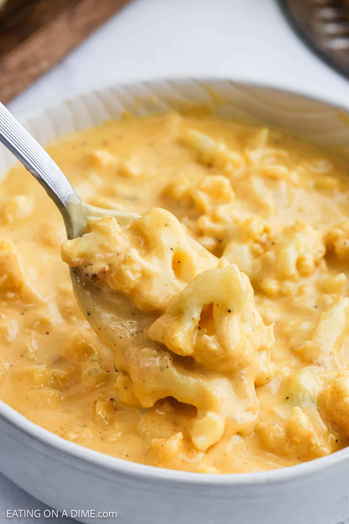 A close-up of a spoonful of creamy, cheesy Cauliflower Mac and Cheese being lifted from a white bowl filled with the same cheesy dish.