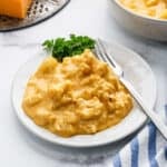 A plate of creamy, cheesy Cauliflower Mac and Cheese casserole garnished with parsley, with a fork resting on the side and a striped napkin nearby.
