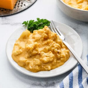 A plate of creamy, cheesy Cauliflower Mac and Cheese casserole garnished with parsley, with a fork resting on the side and a striped napkin nearby.