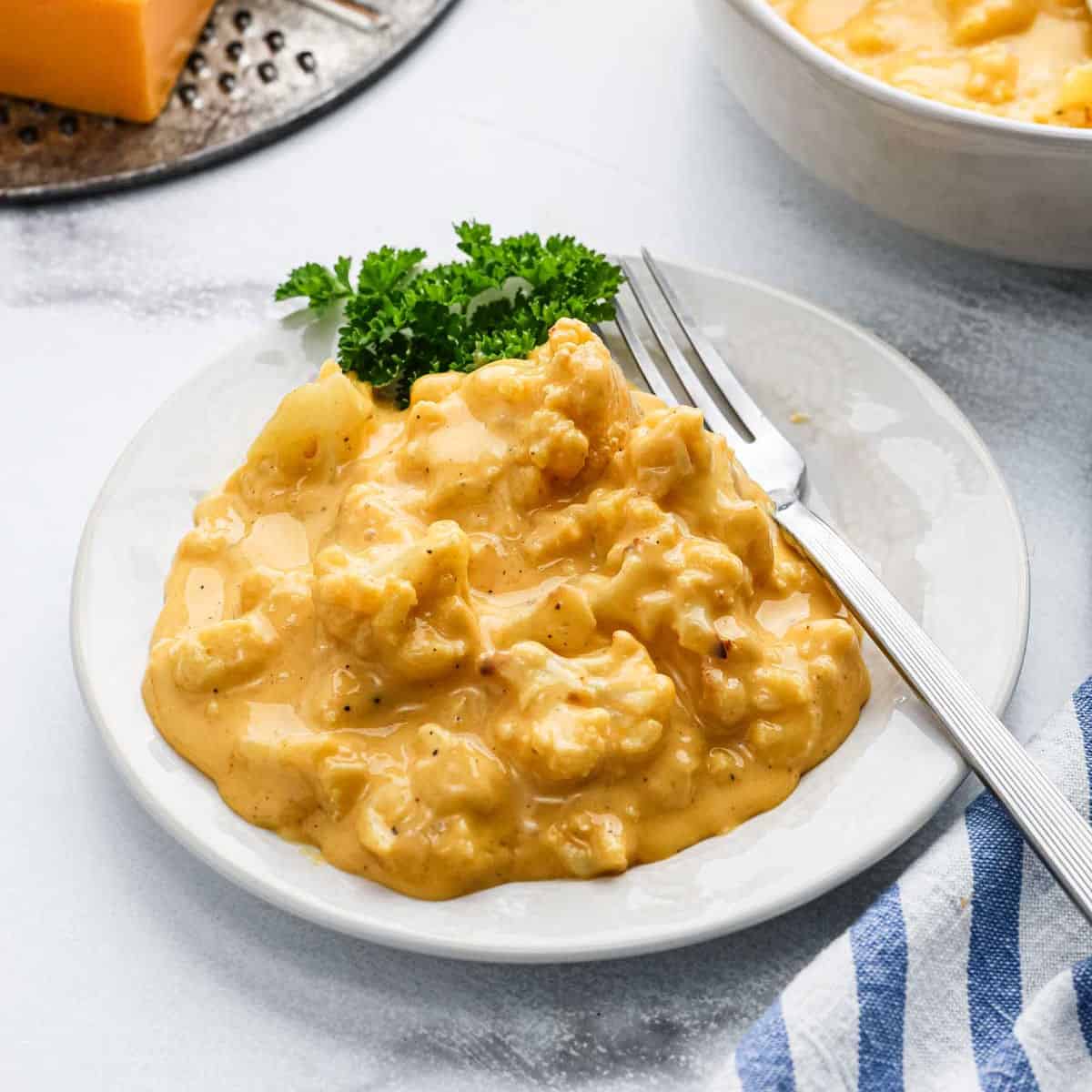 A plate of creamy, cheesy Cauliflower Mac and Cheese casserole garnished with parsley, with a fork resting on the side and a striped napkin nearby.