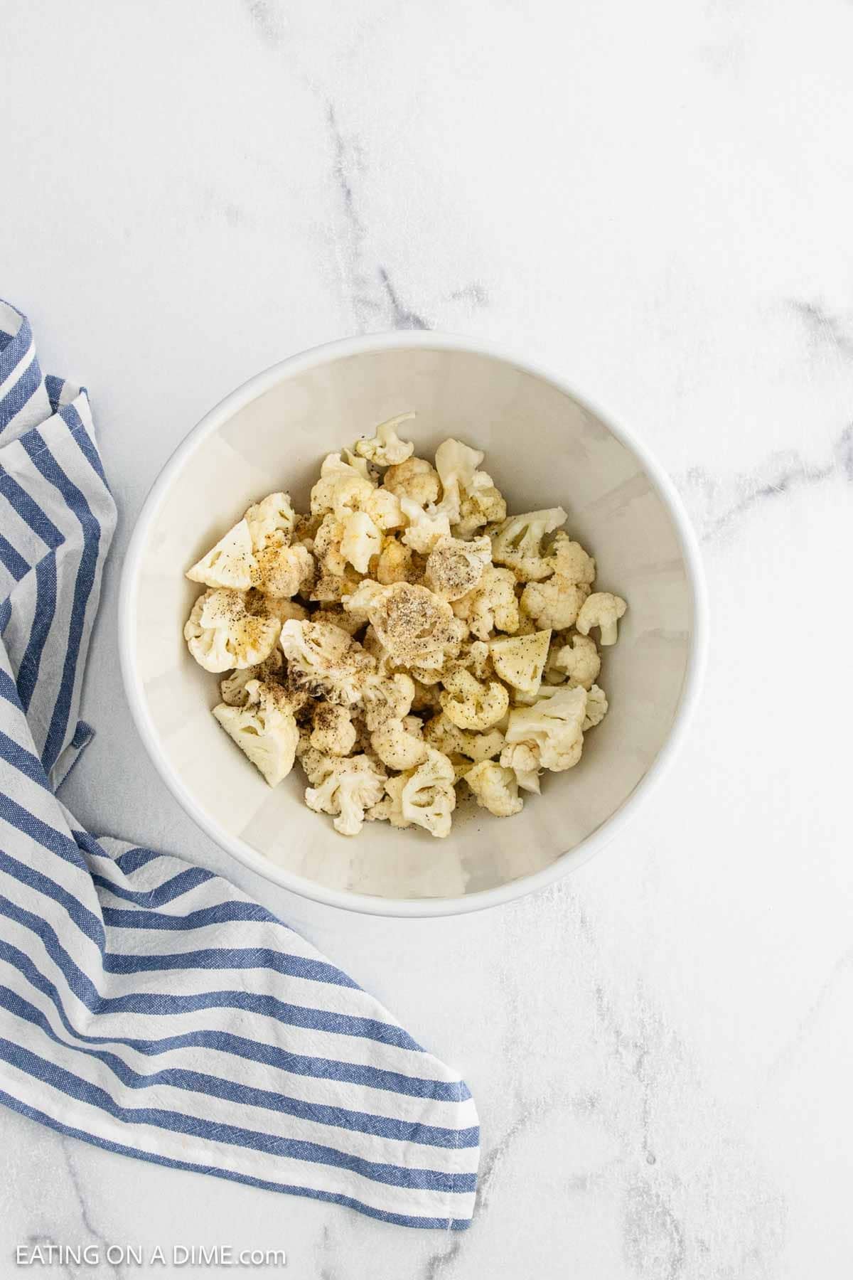 A white bowl filled with spiced cauliflower florets sits on a white surface next to a blue and white striped cloth, evoking the comfort of Cauliflower Mac and Cheese.