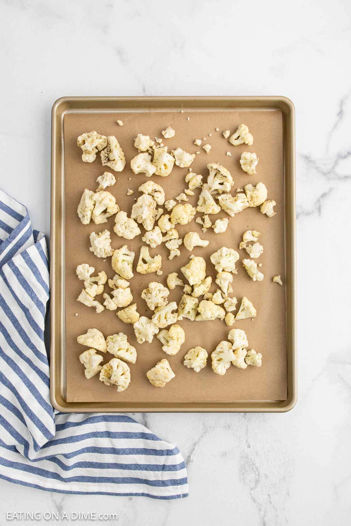A baking sheet lined with parchment paper holds uncooked cauliflower florets, ready to be roasted for a delicious cauliflower mac and cheese, next to a blue and white striped kitchen towel on a white marble surface.