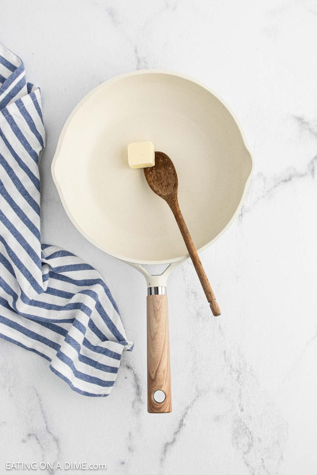 A white frying pan with a wooden handle holds a wooden spoon and a cube of butterโperfect ingredients for starting Cauliflower Mac and Cheese. The pan rests on white marble, next to a blue and white striped cloth.