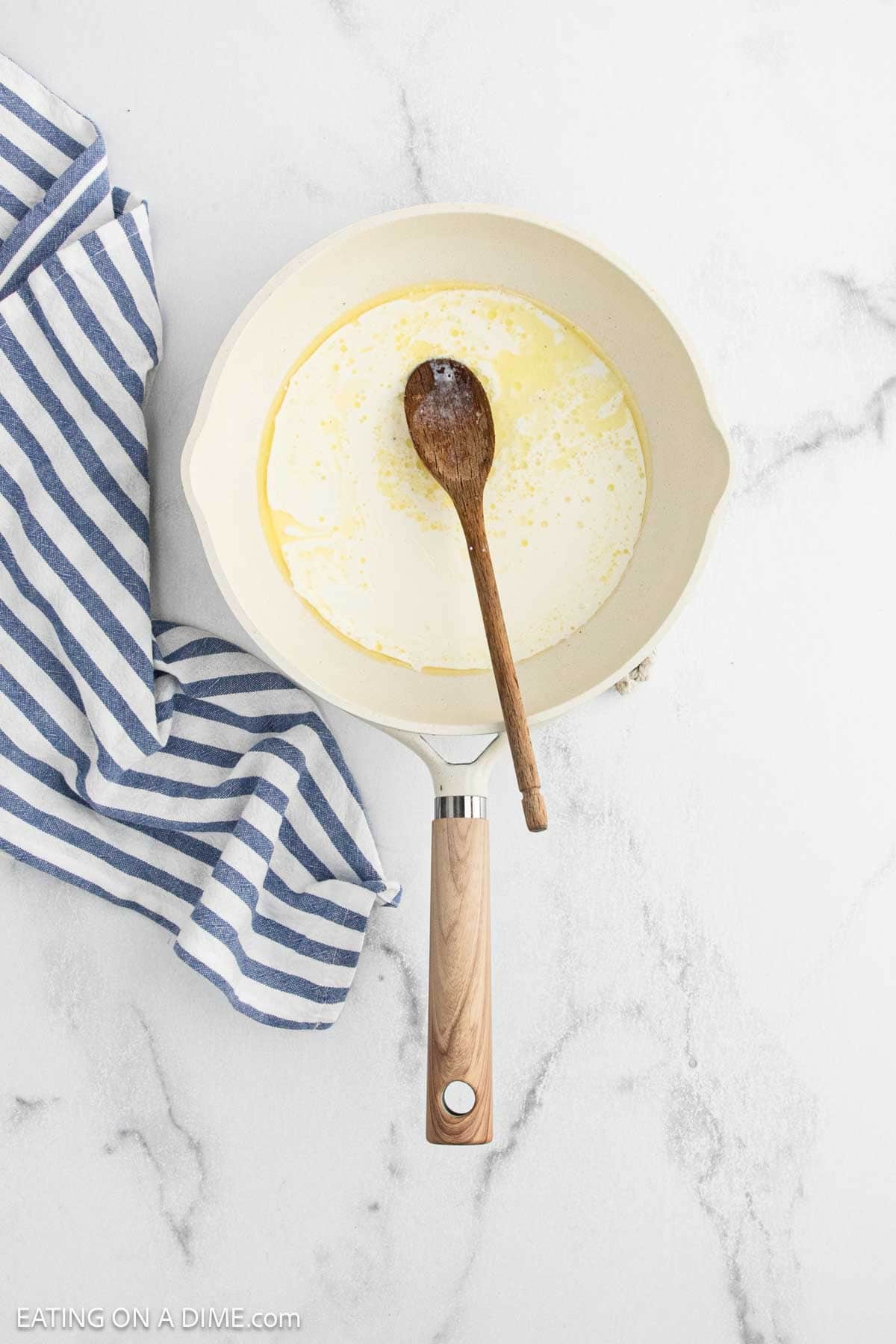 A cream-colored skillet with melted butter and a wooden spoon inside, ready to start Cauliflower Mac and Cheese, sits on a white marble surface next to a blue-and-white striped kitchen towel.