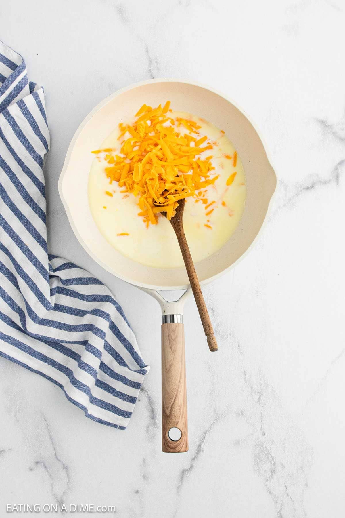 A white skillet with shredded cheddar cheese and milk being stirred by a wooden spoon, perfect for creamy Cauliflower Mac and Cheese, set on a marble countertop next to a blue and white striped kitchen towel.