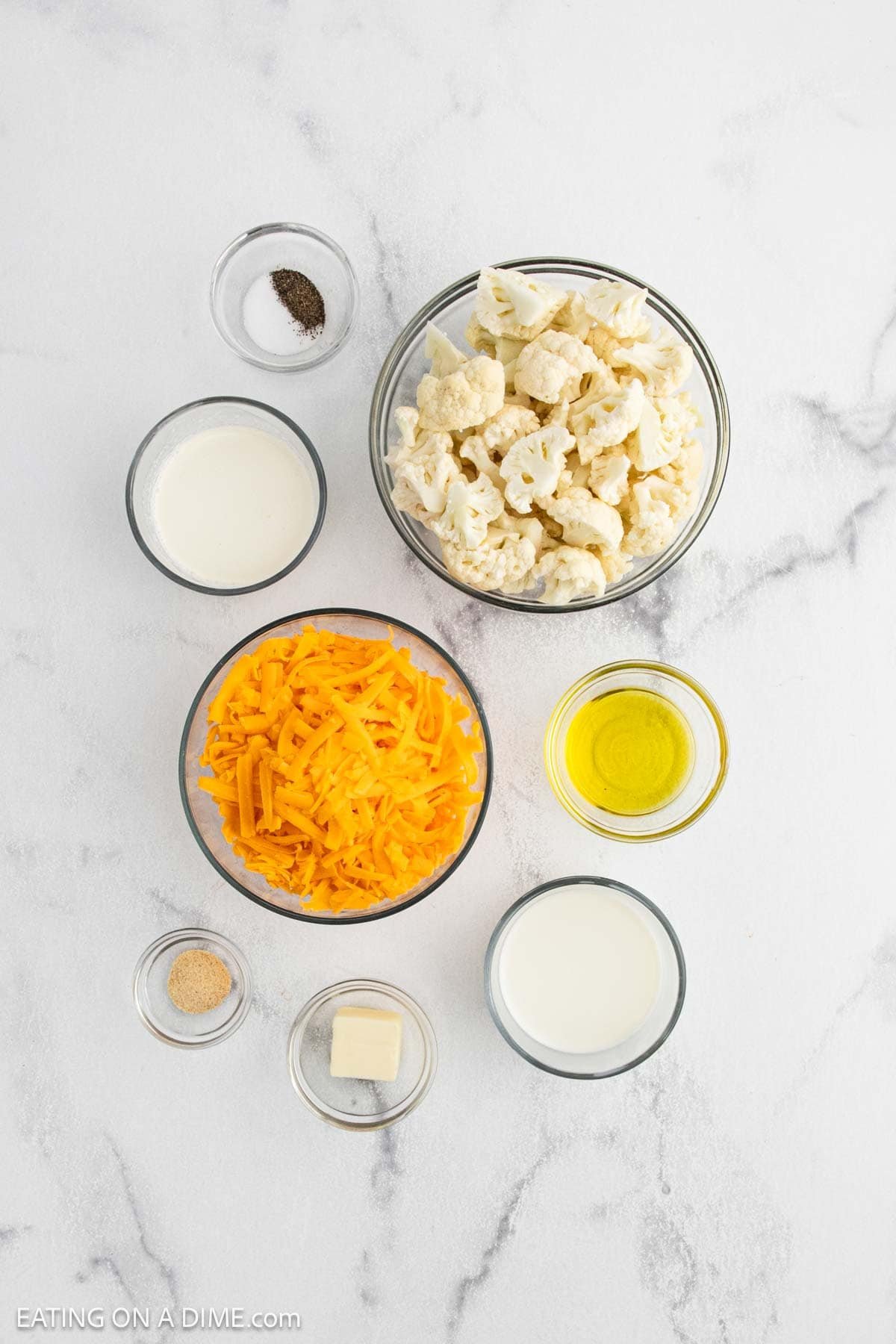 Seven small bowls on a white marble surface contain raw cauliflower florets, shredded cheddar cheese, milk, olive oil, butter, garlic powder, salt, and pepperโperfect for making creamy Cauliflower Mac and Cheese.