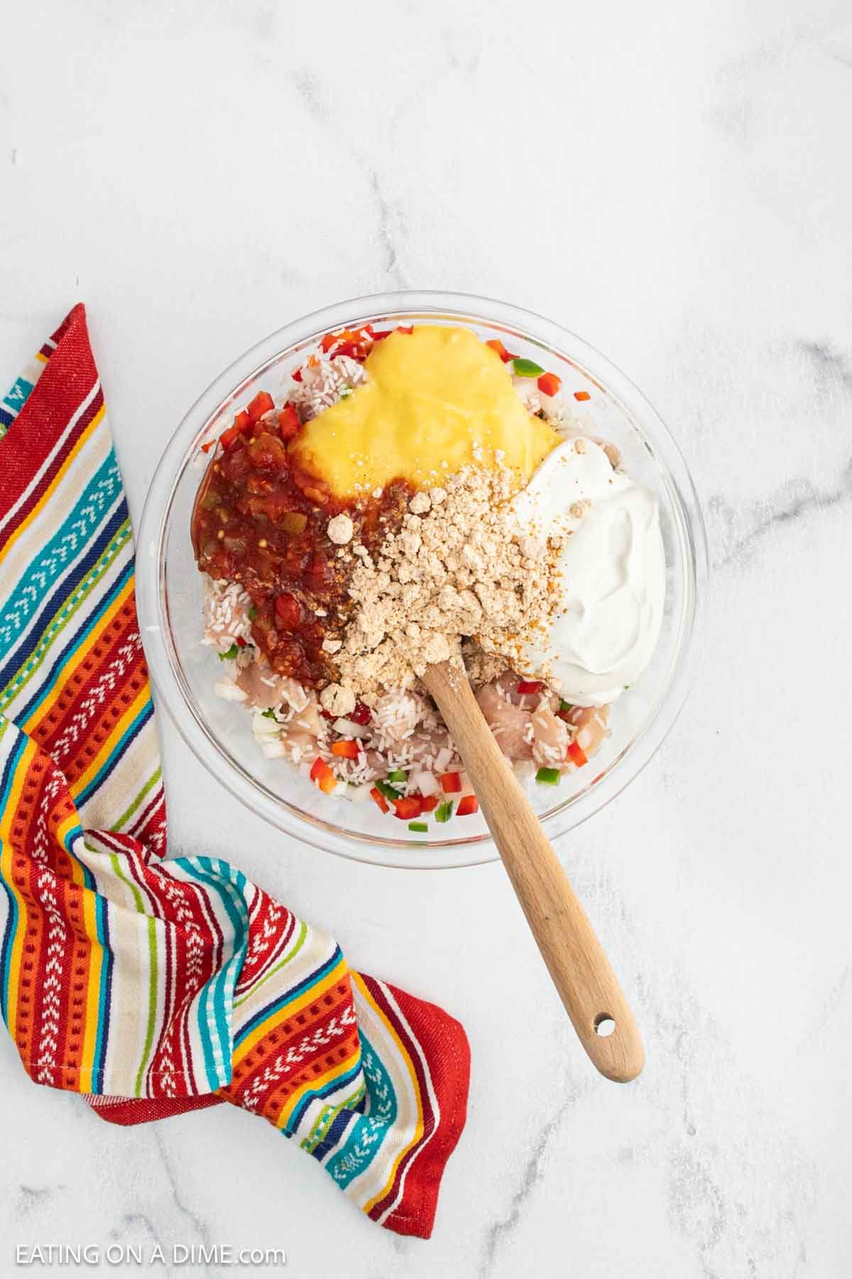 A glass bowl filled with shredded chicken, salsa, sour cream, crushed crackers, and shredded cheese sits on a white marble surface—perfect for a Chicken Fajita Casserole. A wooden spoon rests in the bowl next to a colorful striped cloth.