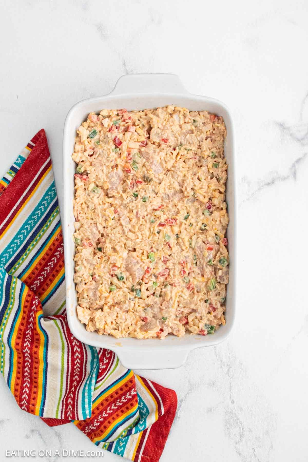 A white casserole dish filled with a creamy Chicken Fajita Casserole, featuring mixed rice and vegetables, sits on a white marble surface next to a colorful striped cloth.