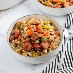 A white bowl filled with rice, topped with sliced sausage, shredded chicken, bell peppers, celery, and okra makes a hearty Meal Plan 82 option. A spoon and a striped cloth napkin rest beside the bowl on a marble surface.