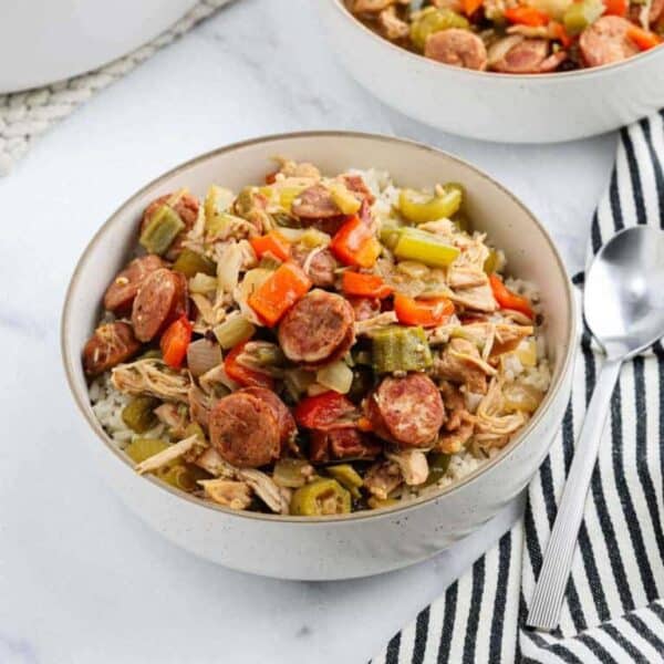 A white bowl filled with rice, topped with sliced sausage, shredded chicken, bell peppers, celery, and okra makes a hearty Meal Plan 82 option. A spoon and a striped cloth napkin rest beside the bowl on a marble surface.