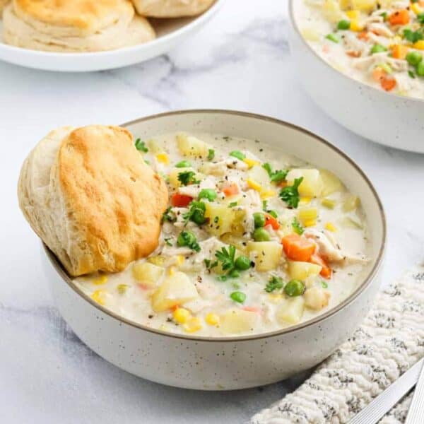 A bowl of creamy chicken and vegetable stew garnished with parsley, served with a biscuit on the side—perfect for Meal Plan 85. A plate of biscuits and another bowl of stew are visible in the background.