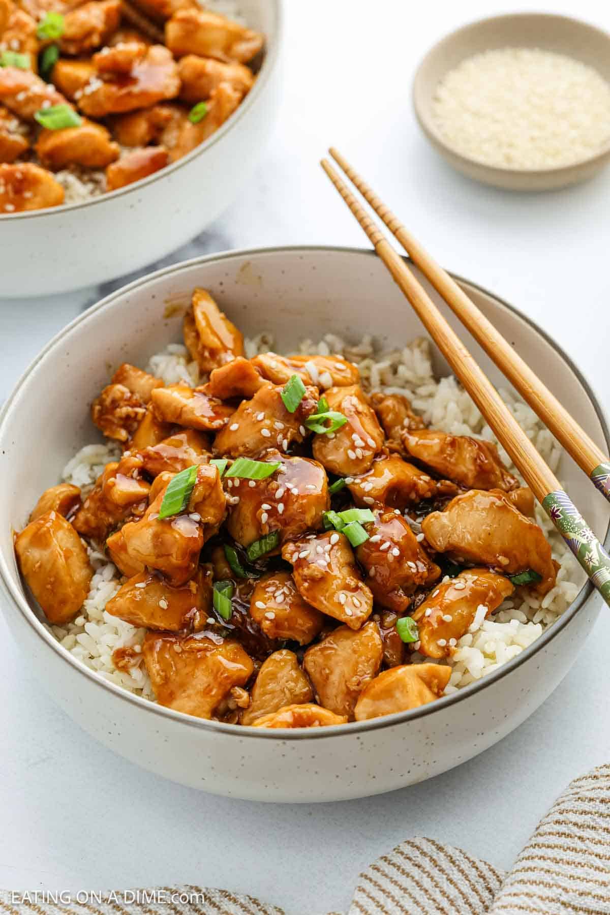 A bowl of white rice topped with glazed Teriyaki Chicken pieces, garnished with chopped green onions and sesame seeds, with wooden chopsticks resting on the bowl. A small dish of sesame seeds is in the background.