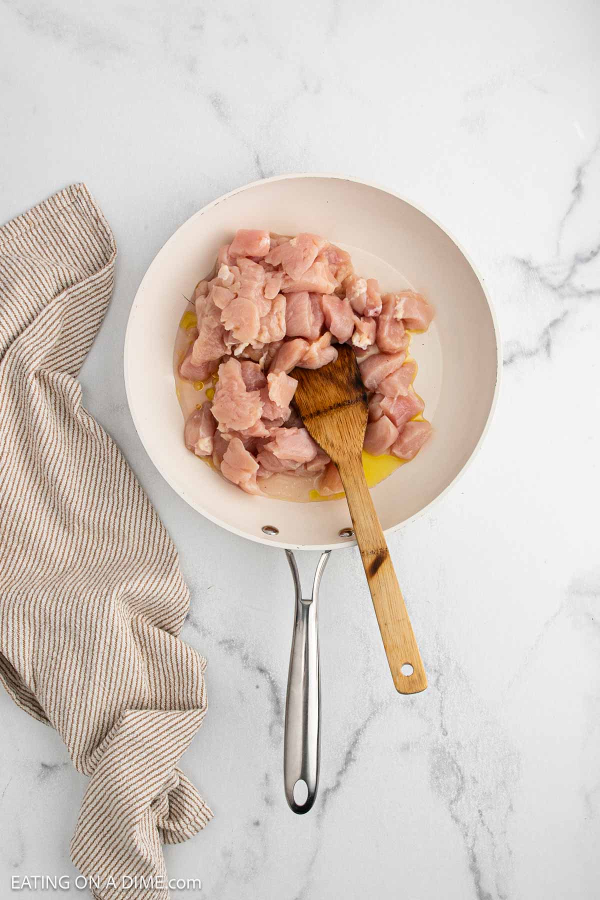A white frying pan with raw, diced chicken pieces—perfect for Japanese Chicken or Chicken Teriyaki—and a wooden spatula sits on a marble countertop next to a beige striped kitchen towel.