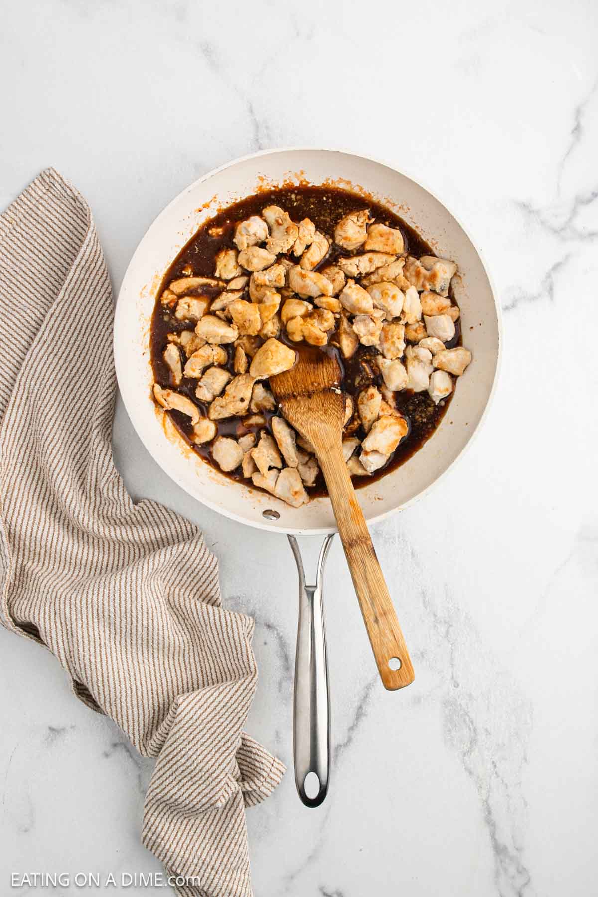 A skillet filled with tender Teriyaki Chicken in a rich brown sauce, with a wooden spatula resting in the pan. A beige striped kitchen towel is draped beside the skillet on a white marble surface.