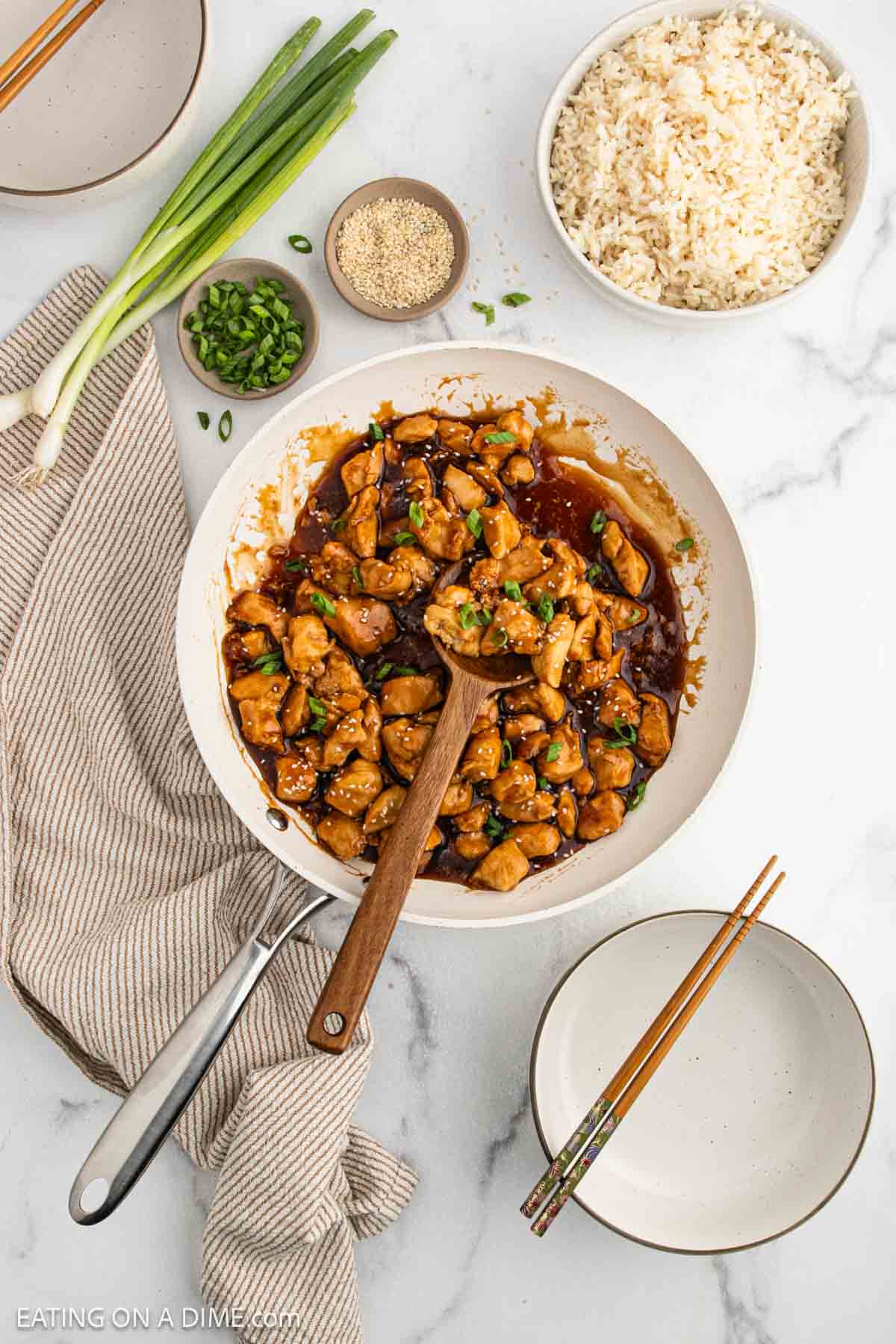 A bowl of Teriyaki Chicken in a rich brown sauce, garnished with green onions, sits on a striped towel beside bowls of cooked rice, sesame seeds, chopped green onions, and two empty bowls with chopsticks.