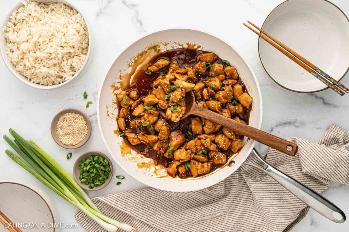 A skillet of flavorful Teriyaki Chicken in a rich brown sauce, garnished with green onions, sits on a striped towel. Nearby are bowls of fluffy white rice, sesame seeds, sliced green onions, plates, and chopsticks on a marble surface.