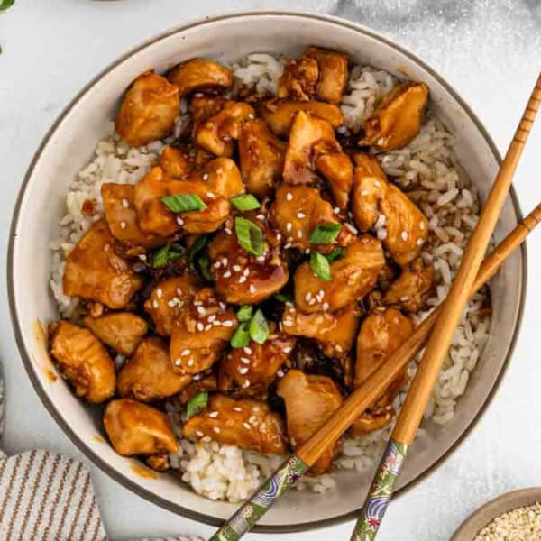 A bowl of rice topped with savory Chicken Teriyaki pieces, glazed to perfection and garnished with sesame seeds and chopped green onions, with a pair of chopsticks resting on the bowl.