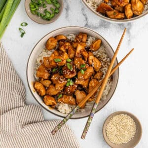 A bowl of rice topped with glazed Teriyaki Chicken pieces, garnished with sesame seeds and chopped green onions. Chopsticks rest on the bowl, with small dishes of sesame seeds and green onions nearby.