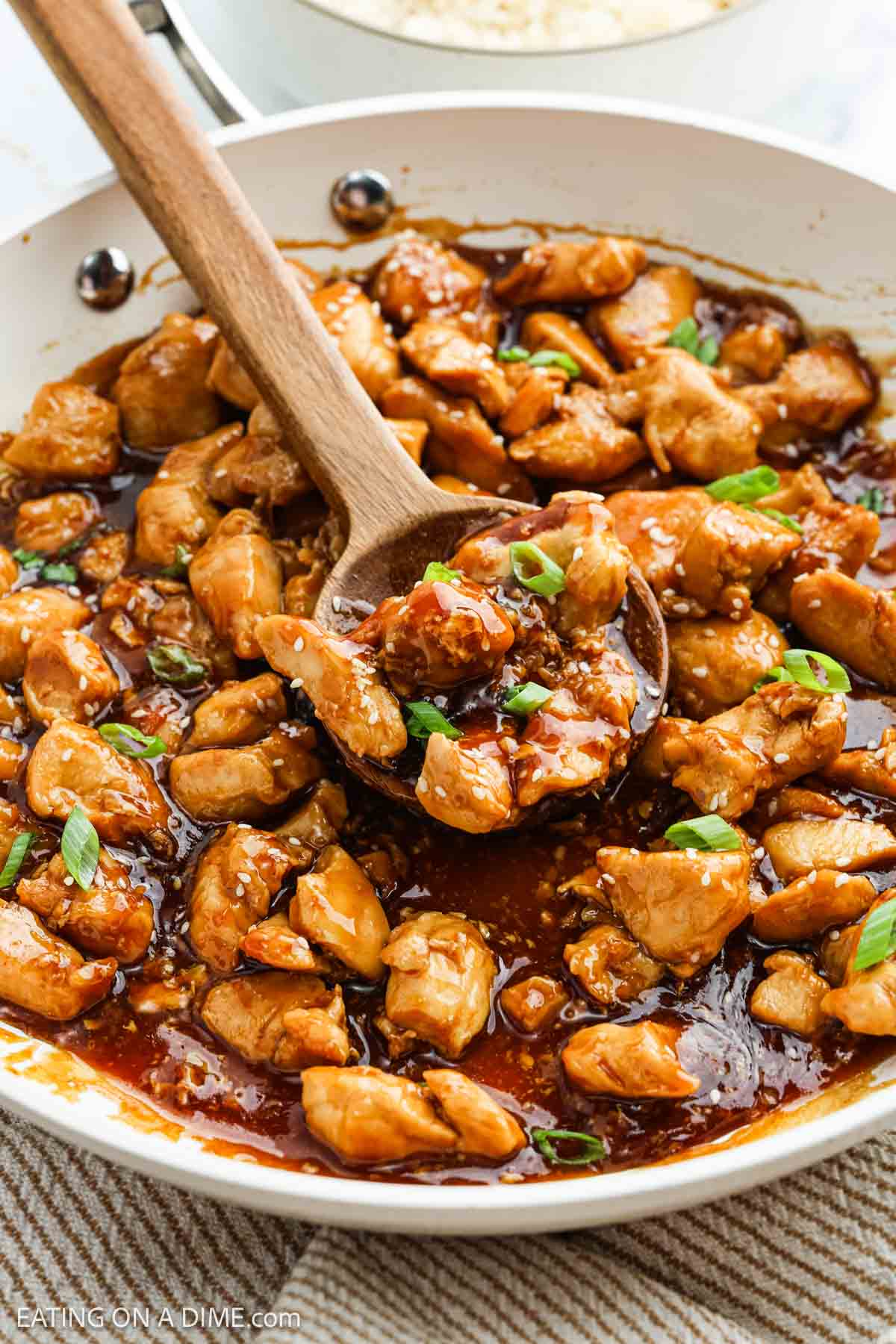 A white skillet filled with glazed Chicken Teriyaki pieces garnished with chopped green onions, with a wooden spoon scooping some of the chicken.