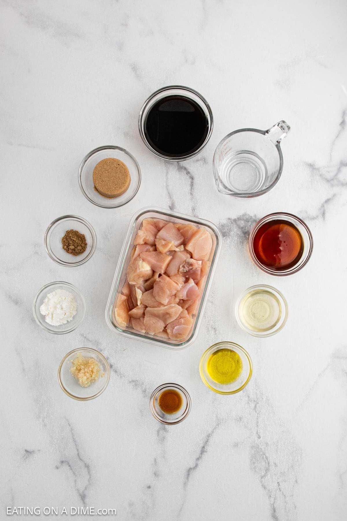 Overhead view of raw cubed chicken in a glass container surrounded by small bowls of ingredients for Japanese Chicken Teriyaki, including soy sauce, brown sugar, water, oil, cornstarch, garlic, and spices on a white surface.