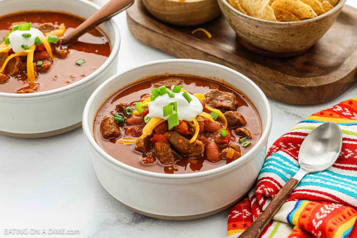 Two bowls of Chili Con Carne topped with sour cream, shredded cheese, and chopped green onions sit on a table. A bowl of tortilla chips, a colorful napkin, and a spoon are nearby.