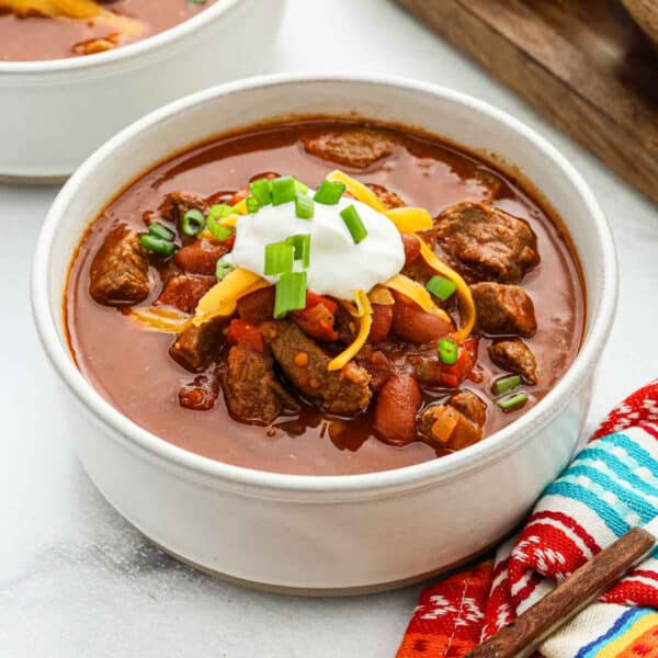 A bowl of hearty Chili Con Carne topped with sour cream, shredded cheddar cheese, and chopped green onions sits beside a colorful napkin and a wooden spoon.