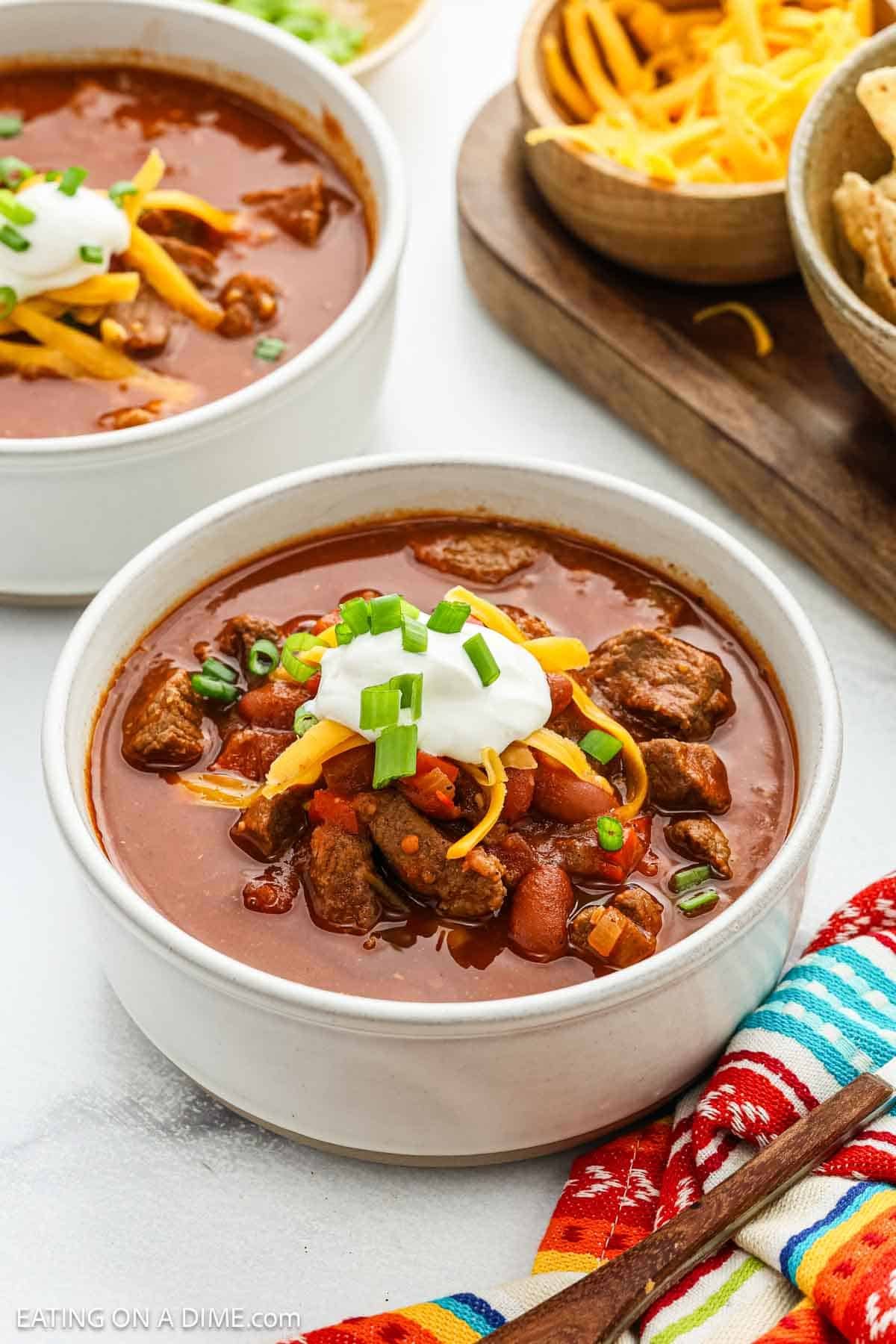 A bowl of Chili Con Carne topped with shredded cheese, sour cream, and chopped green onions sits on a table next to a colorful napkin, with another bowl of chili, grated cheese, and tortilla chips in the background.