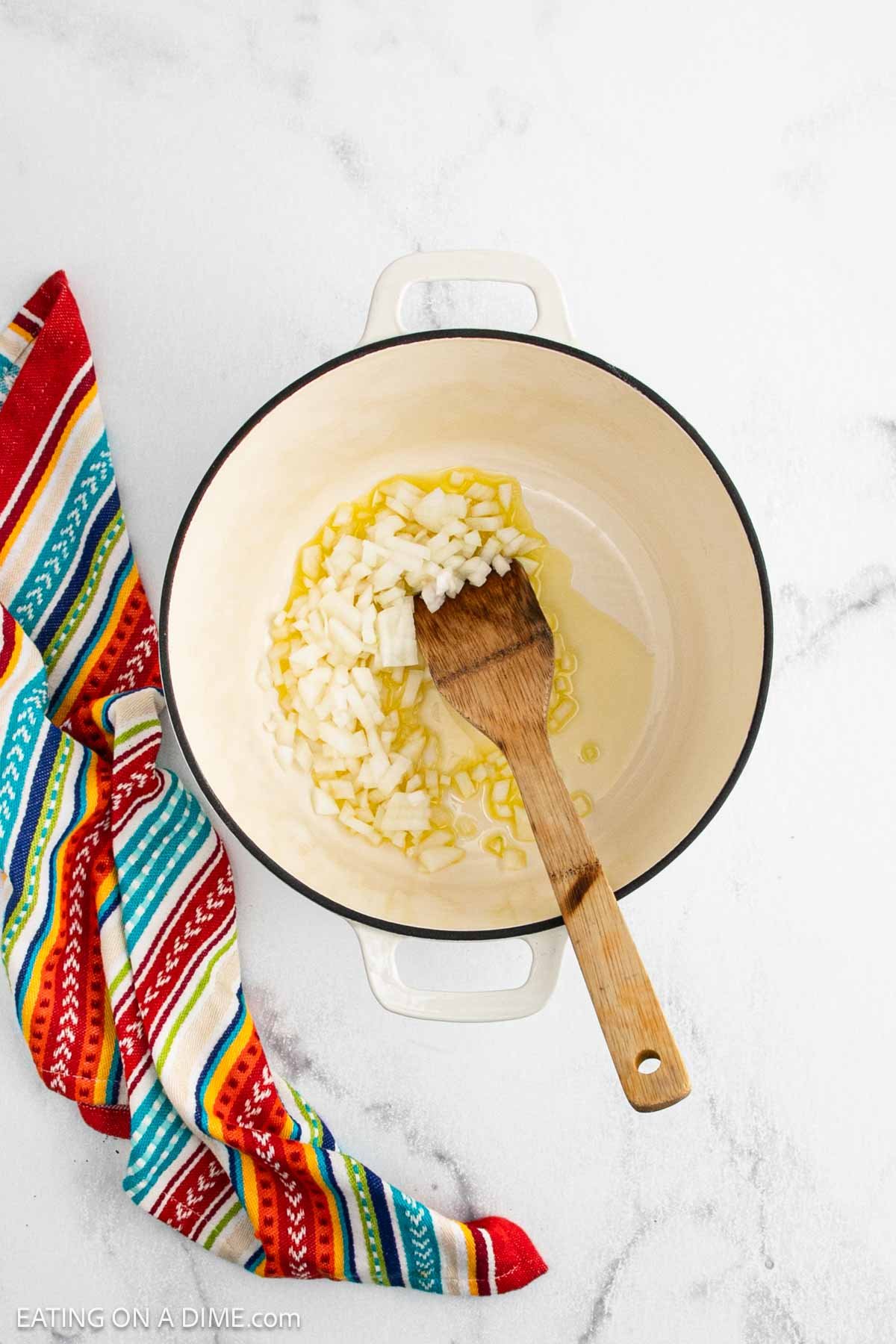 A white pot with chopped onions and oil, the savory base for Chili Con Carne, sits on a light countertop. A wooden spatula rests in the pot, with a colorful striped kitchen towel nearby.