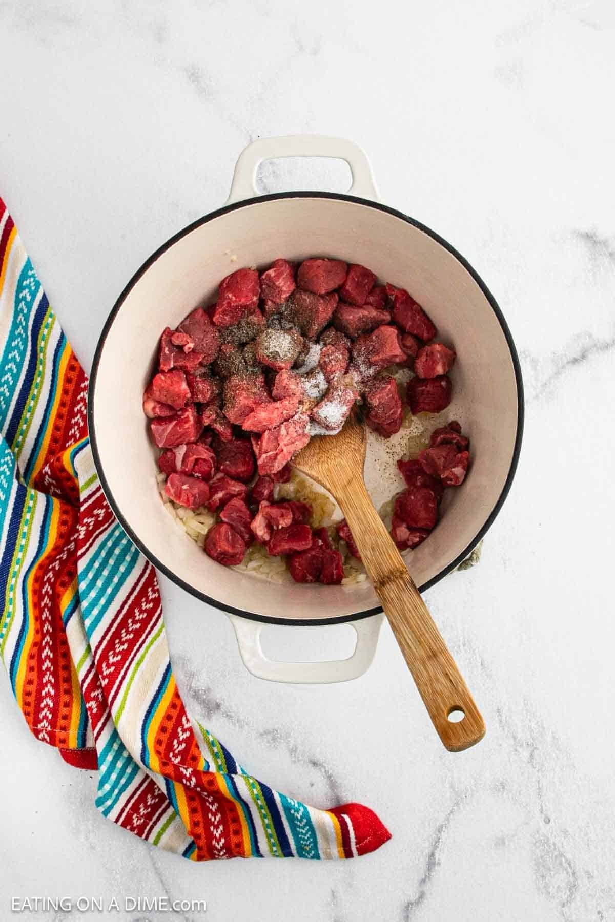 Chunks of raw beef with seasonings in a white Dutch oven, being stirred with a wooden spoon—perfect beginnings for Chili Con Carne. A colorful striped kitchen towel is draped beside the pot on a white countertop.