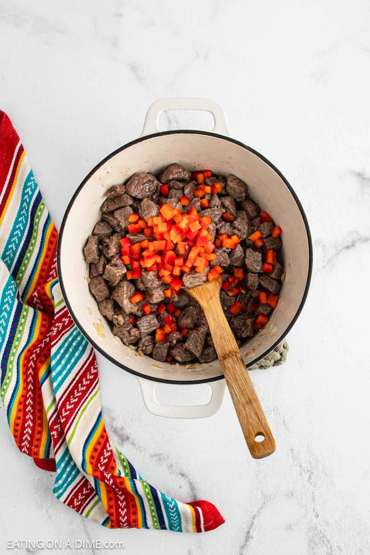 A white pot with cooked beef cubes and chopped red bell peppers, stirred with a wooden spoon, brings Chili Con Carne to life. A colorful striped cloth sits beside the pot on a light marble surface.