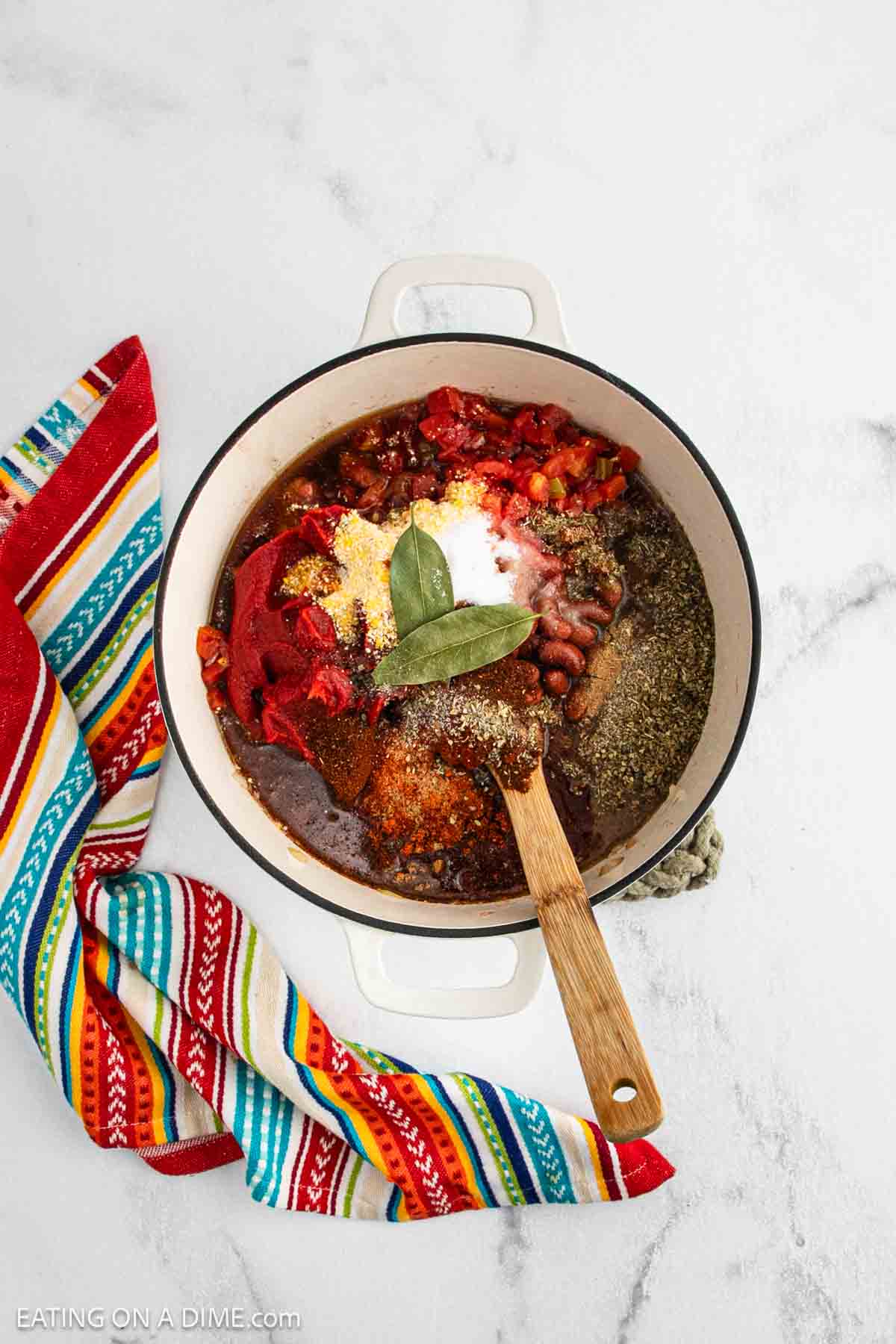 A white pot filled with Chili Con Carne ingredients—beans, tomatoes, spices, and bay leaves—sits on a white surface next to a colorful striped kitchen towel. A wooden spoon rests inside the pot.