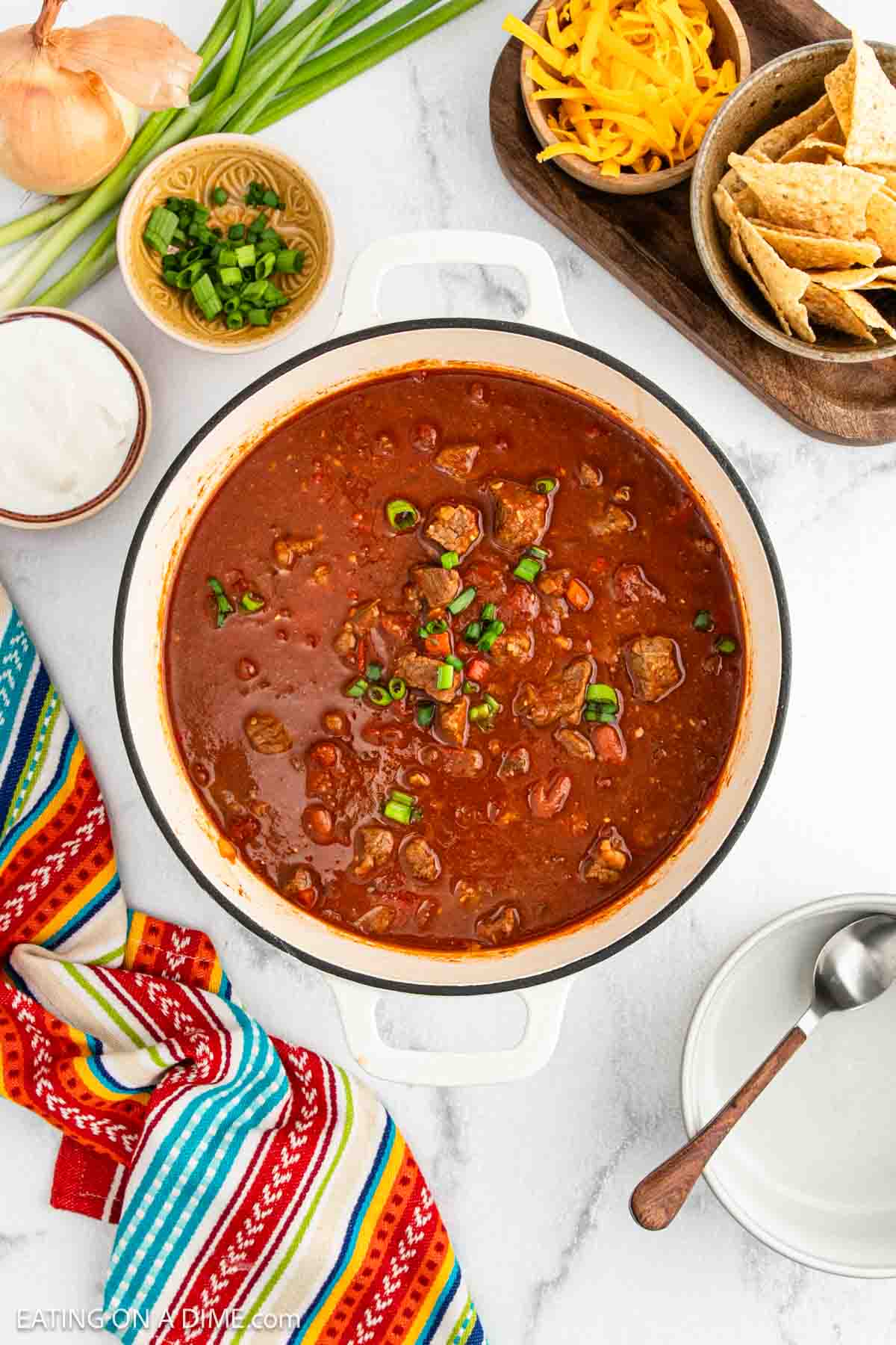 A pot of chunky Chili Con Carne with beans and beef, garnished with chopped green onions, sits on a marble counter next to shredded cheese, tortilla chips, sour cream, and fresh vegetables. A colorful striped napkin is nearby.