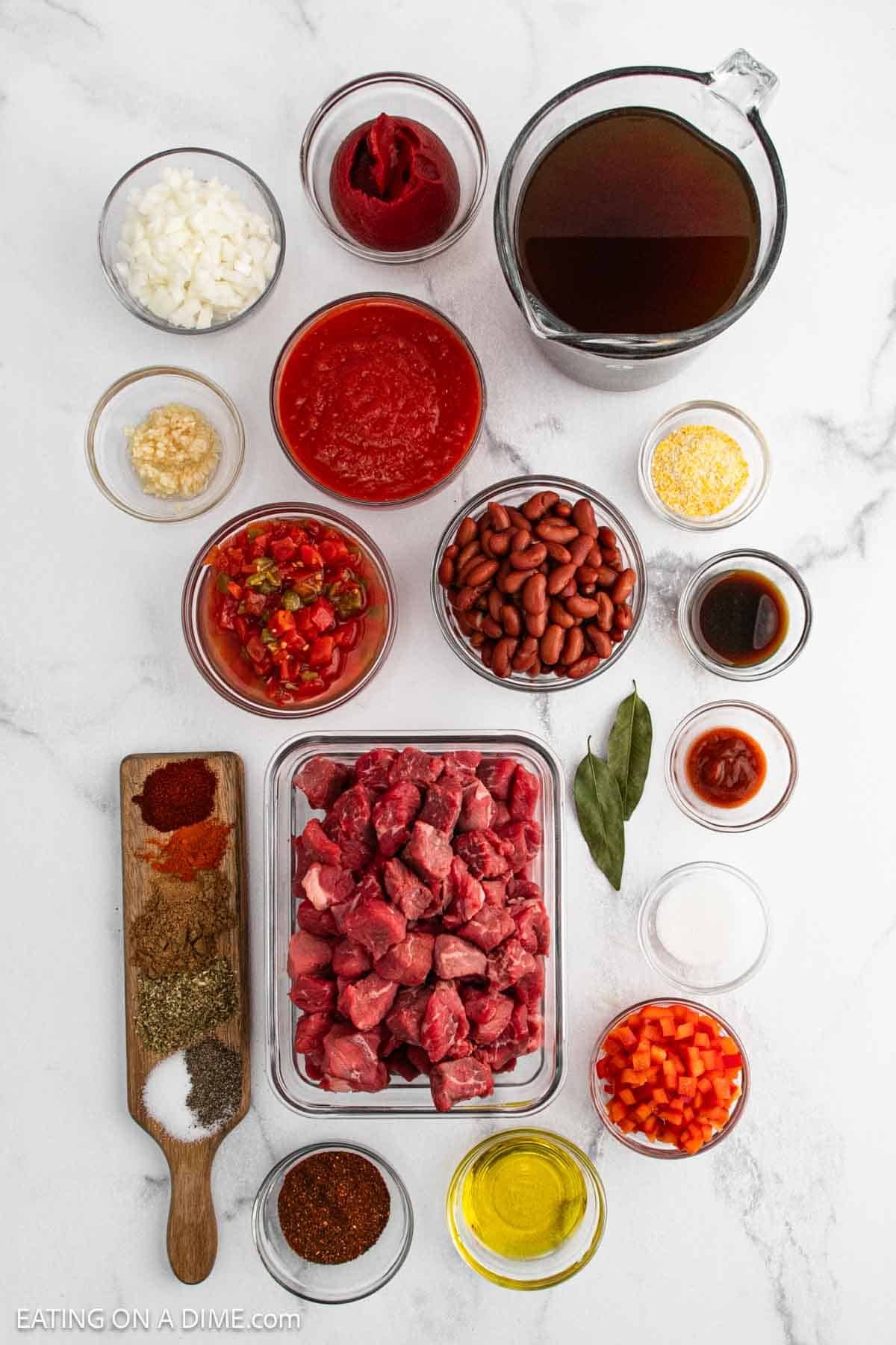 Overhead view of Chili Con Carne ingredients on a white surface, including diced beef, kidney beans, chopped onions, diced tomatoes, spices, crushed tomatoes, broth, garlic, bell pepper, tomato paste, oil, and bay leaves in bowls and containers.