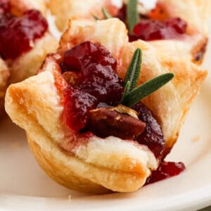 A close-up of Cranberry Brie Bites—puff pastry cups filled with cranberry sauce, a nut, and garnished with a fresh sprig of rosemary—served on a white plate.