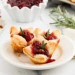 Three Brie bites filled with cranberry sauce and garnished with rosemary are arranged on a small white plate. A dish of cranberry sauce and a sprig of rosemary are visible in the background.