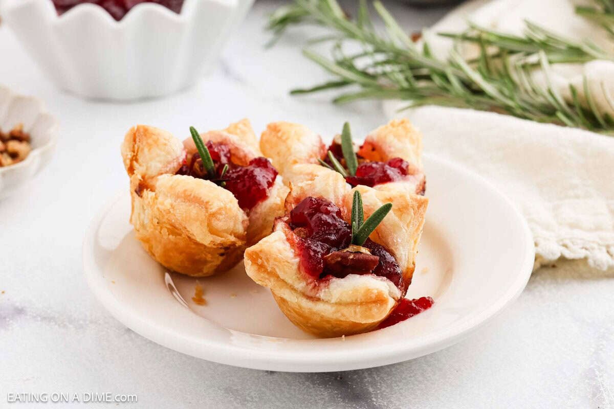 Three Cranberry Brie Bites, perfect as a holiday appetizer, are filled with cranberry sauce and pecans, then garnished with rosemary sprigs on a white plate. A bowl of cranberry sauce and fresh rosemary are visible in the background.