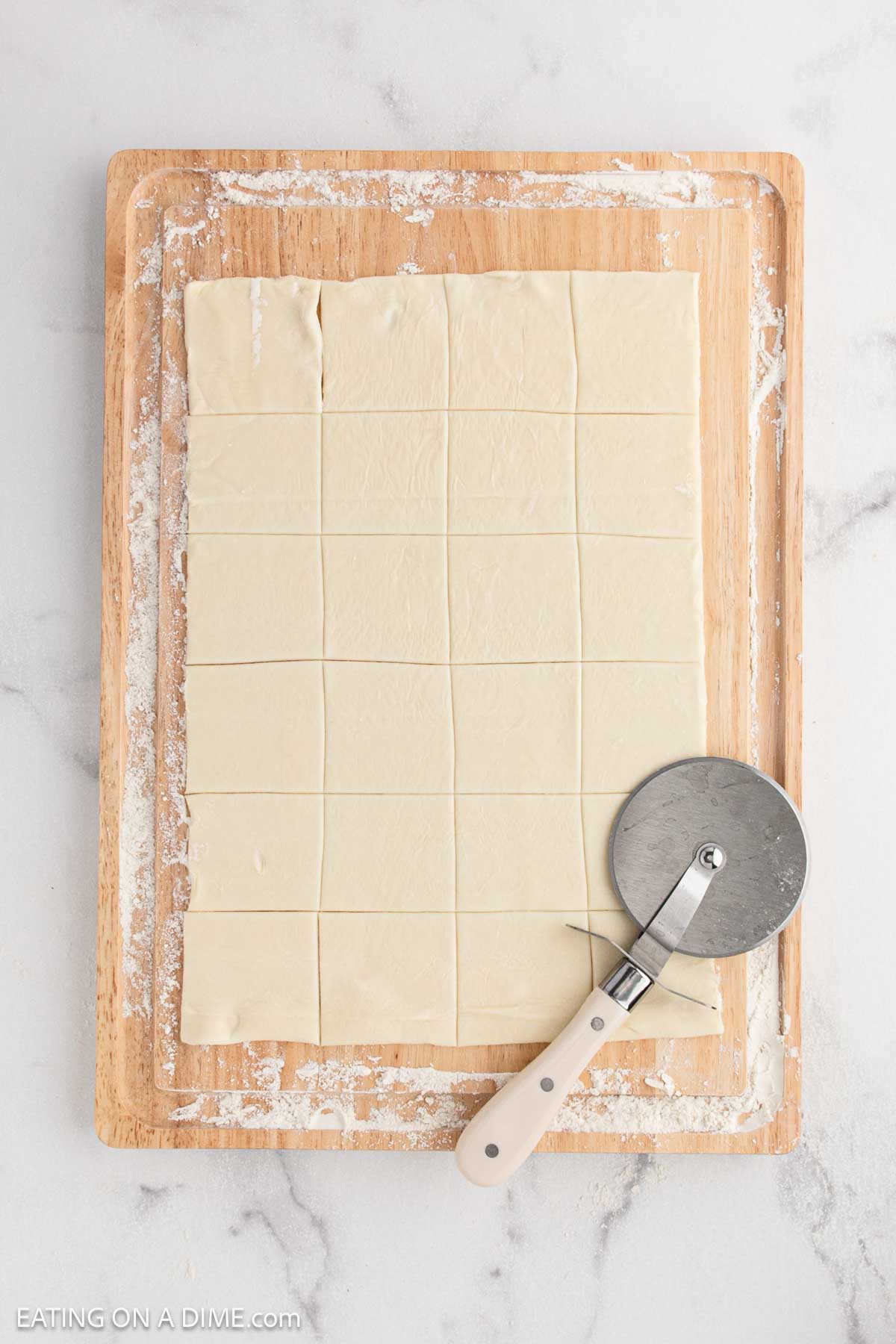 A wooden cutting board with a sheet of dough cut into squares, perfect for making cranberry brie bites, and a pizza cutter resting on the board. The surface is lightly dusted with flour.