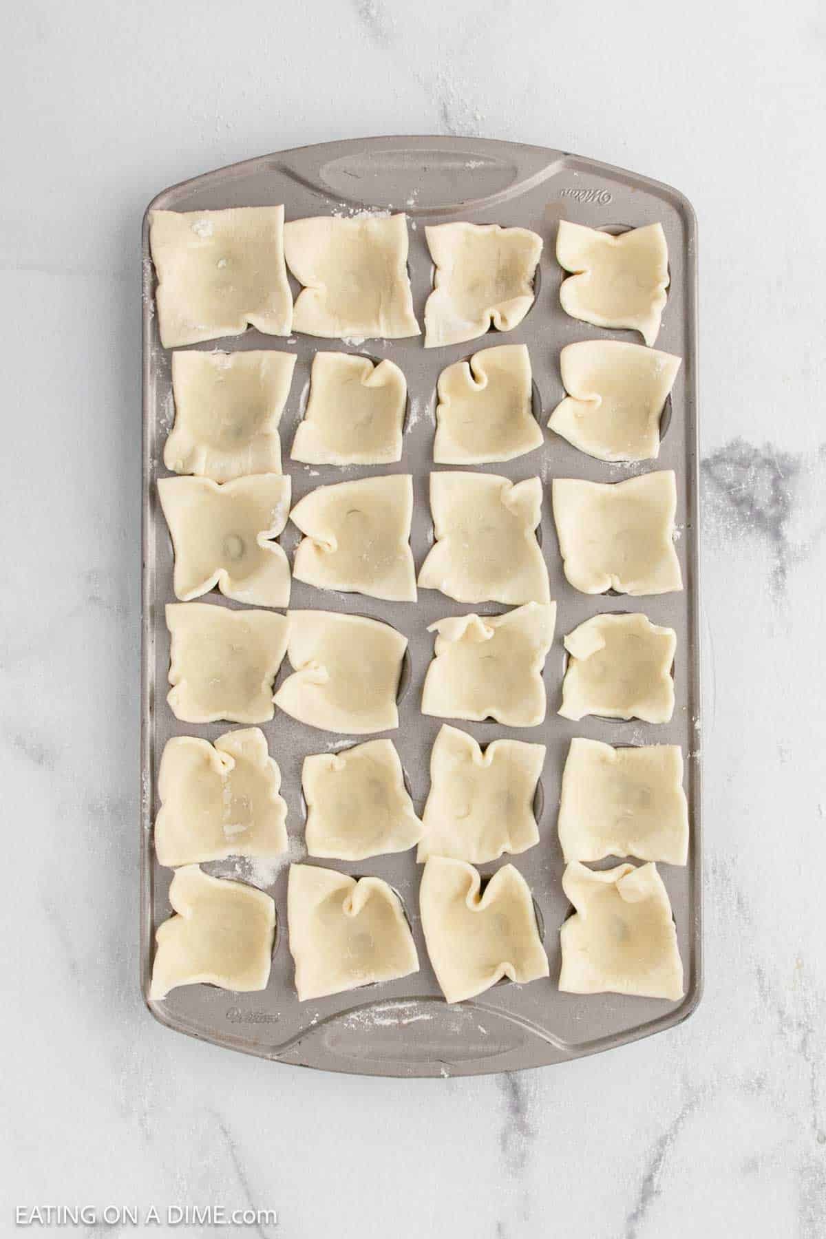 A baking sheet with 24 uncooked, square-shaped dough pieces, each folded and pinched in the center to make perfect Brie bites, arranged in neat rows on a light marble surface.