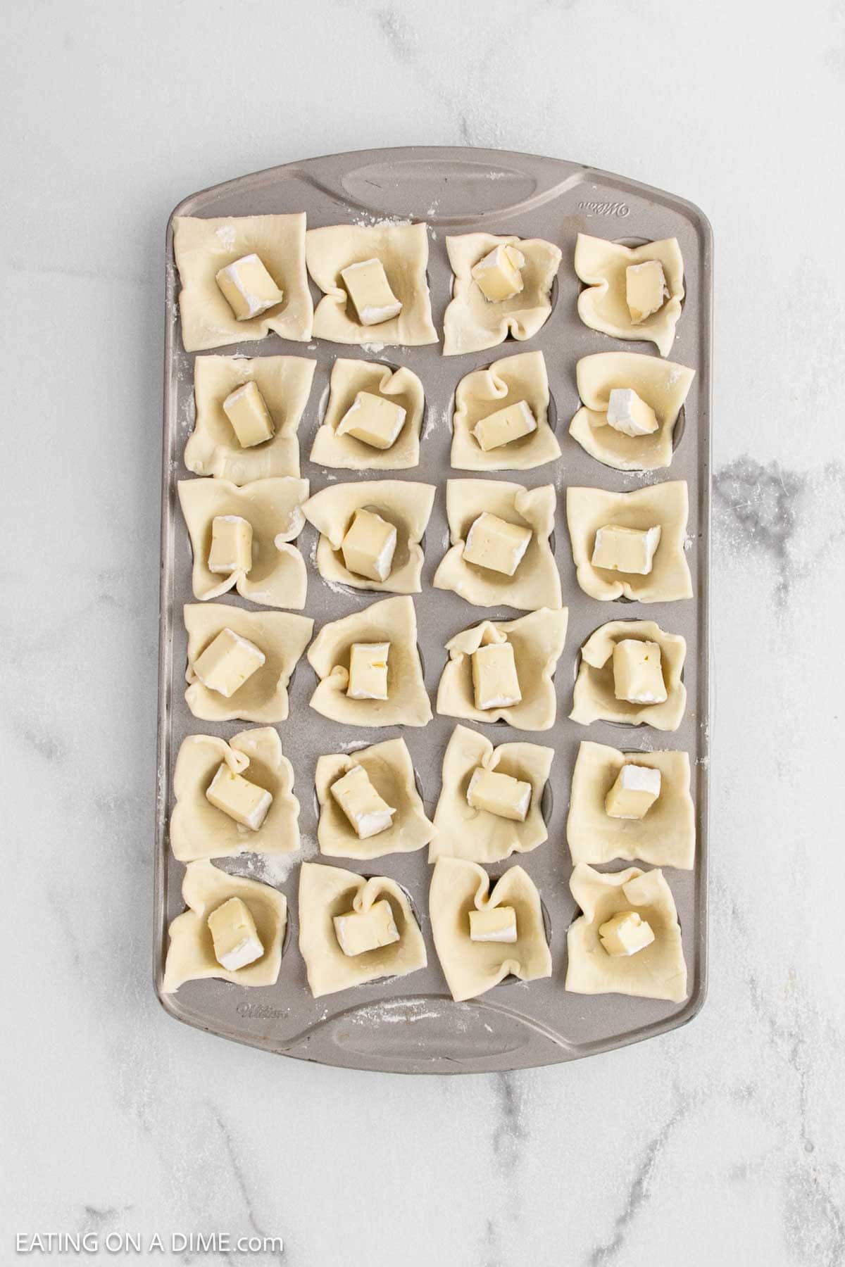 A mini muffin tin filled with squares of dough, each holding a small piece of brie cheese and cranberry, ready to be baked into delicious Brie Bites on a light marble surface.