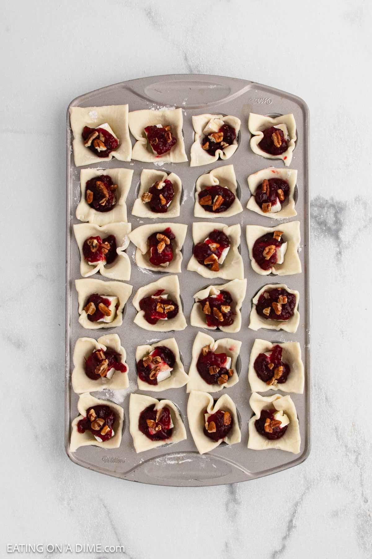 A baking tray with 24 mini muffin cups, each lined with pastry dough and filled with cranberry, brie, and chopped nuts, ready to become delicious bites. The tray sits on a light marble surface.