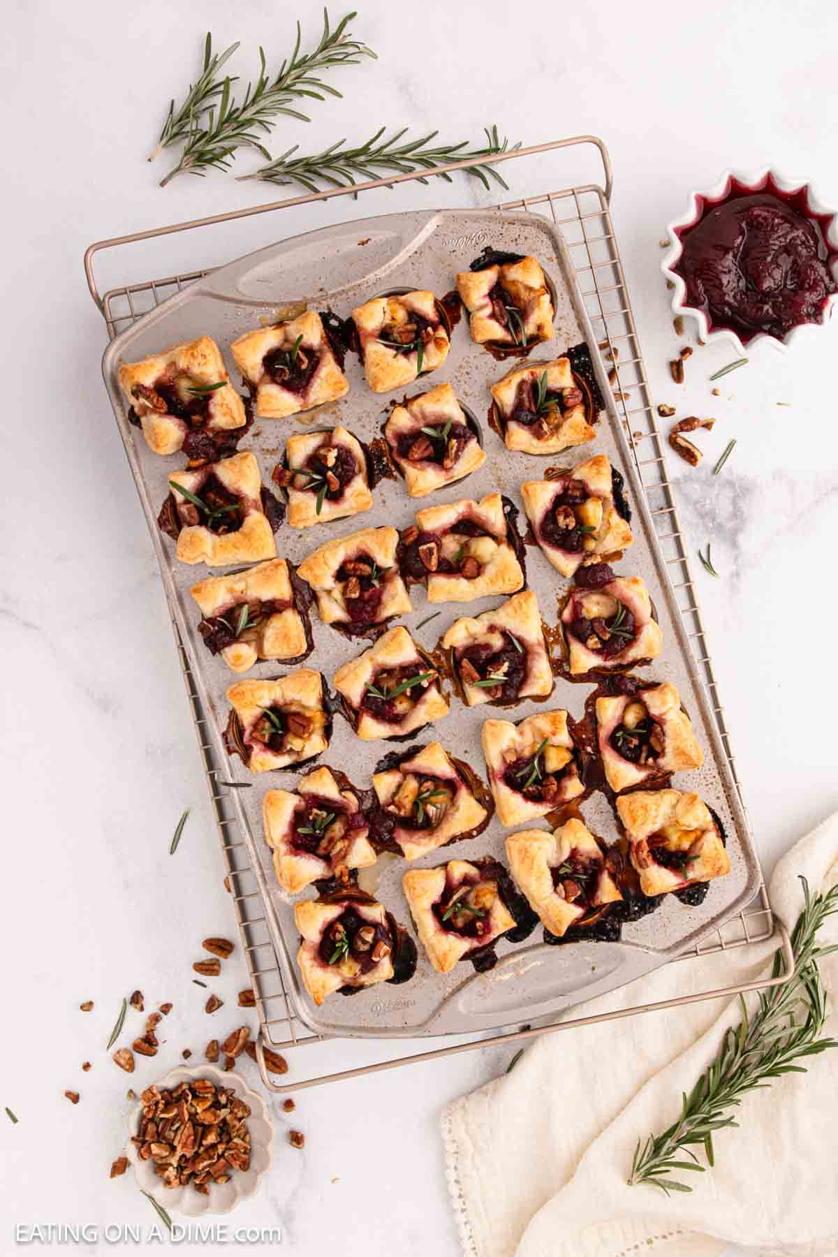 A baking sheet with 24 Brie Bites, topped with cranberry sauce, nuts, and herbs, sits on a cooling rack. This festive appetizer is surrounded by a bowl of cranberry sauce, pecans, rosemary sprigs, and a white napkin.