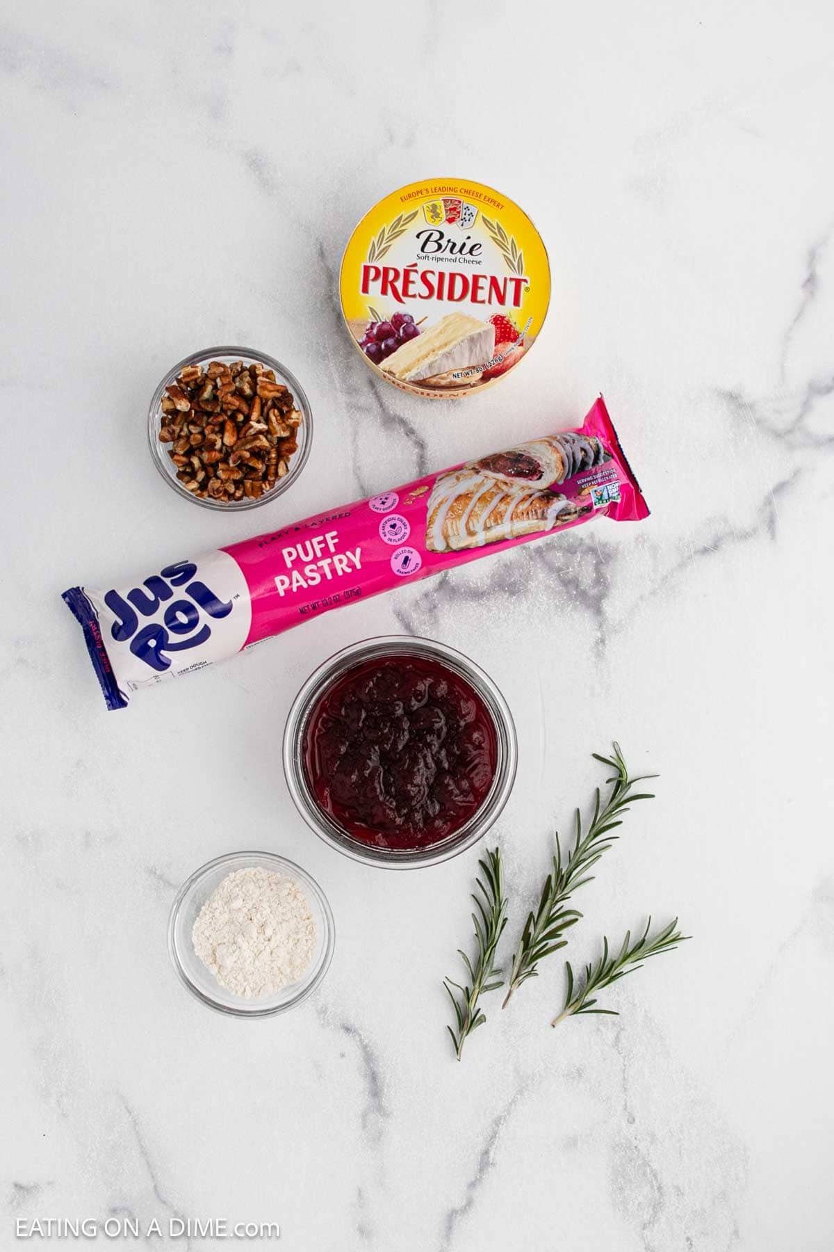 A flat lay of ingredients for Cranberry Brie Bites on a white surface: round Brie cheese, puff pastry, cranberry sauce, chopped pecans, a spoonful of flour, and a sprig of rosemary.