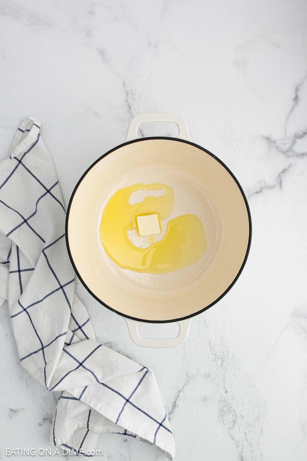 A white Dutch oven with a pat of butter melting inside sits on a white marble surface, ready to start a batch of creamy chicken noodle soup, next to a white kitchen towel with a blue grid pattern.