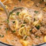 A close-up of a ladle scooping creamy tortellini soup with ground meat, spinach, and herbs from a pot. The creamy soup features a rich, orange-tinted broth with visible chunks of pasta and vegetables.