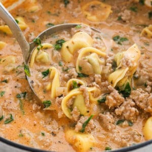 A close-up of a ladle scooping creamy tortellini soup with ground meat, spinach, and herbs from a pot. The creamy soup features a rich, orange-tinted broth with visible chunks of pasta and vegetables.