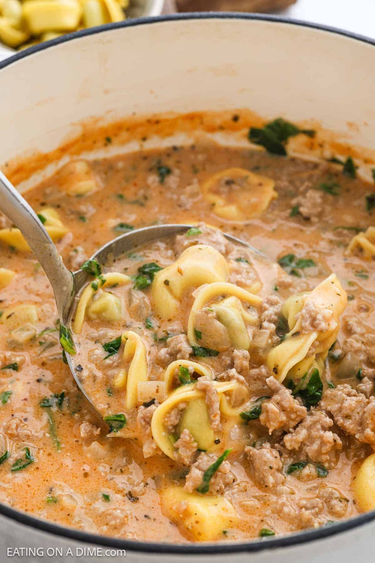 A close-up of a pot filled with Creamy Tortellini Soup, packed with ground meat, spinach, and herbs. A metal ladle scoops up the rich, hearty texture, showcasing the soup’s deliciously creamy appeal.