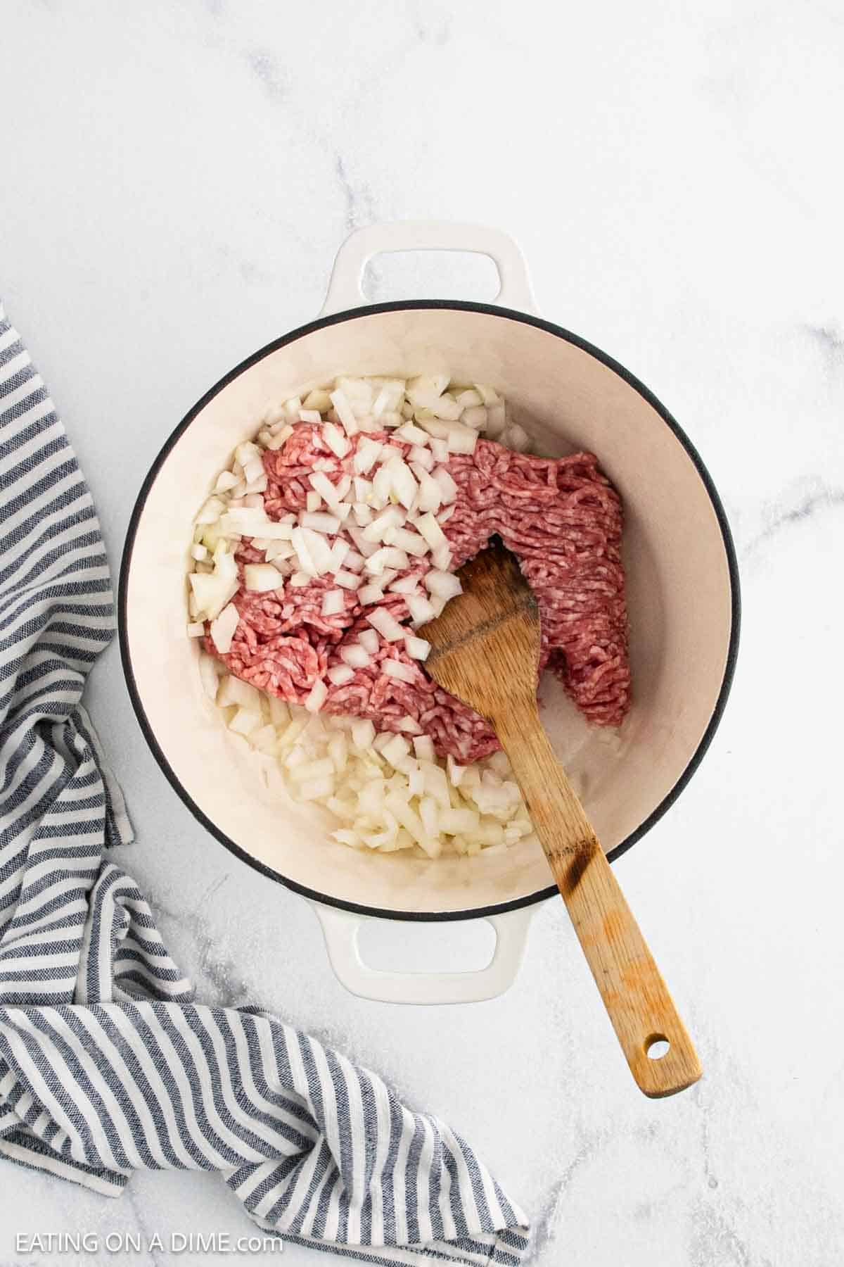 A white pot on a marble surface contains raw ground beef and chopped onions being stirred with a wooden spoon, the first step in making flavorful Creamy Tortellini Soup. A striped kitchen towel sits next to the pot.