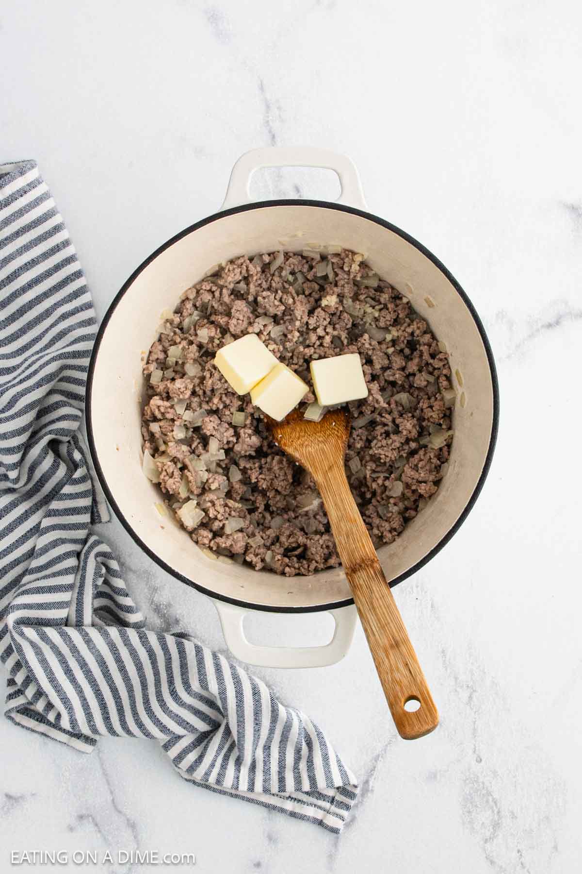 A white pot filled with cooked ground beef and onions, ready for a delicious creamy tortellini soup, with three cubes of butter and a wooden spoon on top. A striped kitchen towel rests beside the pot on a marble countertop.