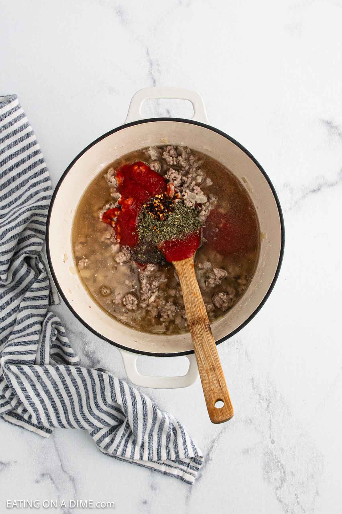 A white pot filled with creamy tortellini soup, featuring cooked ground beef, tomato sauce, spices, and broth, sits on a marble surface. A wooden spoon rests in the pot, with a striped kitchen towel beside it.