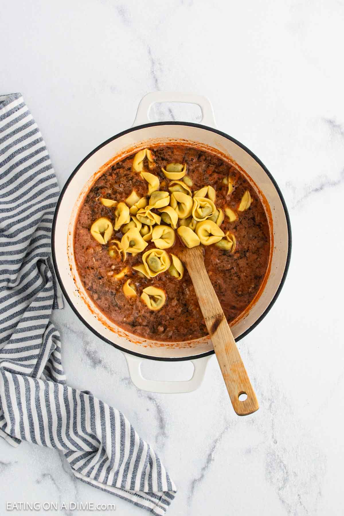 A white pot filled with hearty Tortellini Soup—rich tomato and beef broth, topped with cheese tortellini—rests on a marble surface beside a striped towel. A wooden spoon sits inside, ready to serve this comforting meal.
